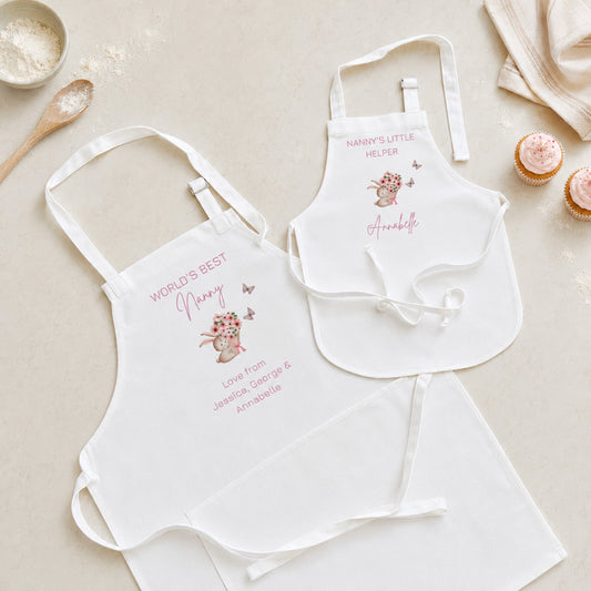 Two white baby bib aprons laid flat on a light beige surface with styled kitchen props. One bib reads "WORLD'S BEST Nanny" with a pink bunny illustration and personalized text below; the other reads "NANNY'S LITTLE MILITER" with a bunny and the name "Annabelle" in pink script.