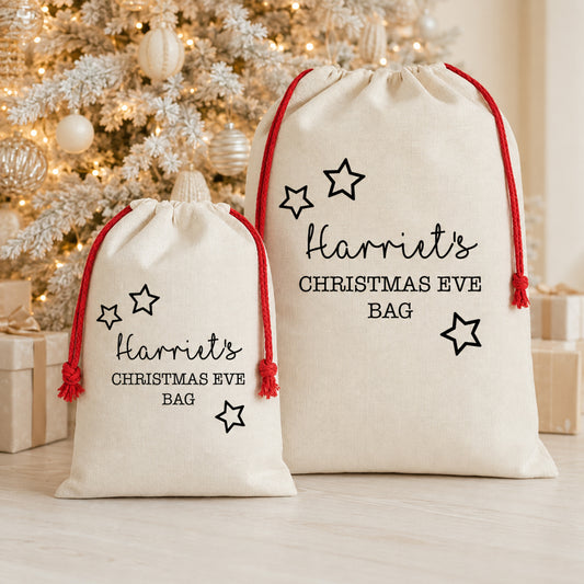 Two personalized cream canvas Christmas Eve bags with red drawstrings and black text reading 'Harriet's CHRISTMAS EVE BAG' with star graphics, displayed in front of a decorated Christmas tree.