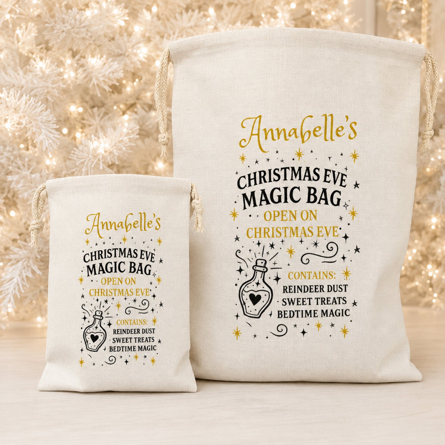 Two cream canvas drawstring bags with gold and black printed holiday text and illustration, shown on a light surface with blurred warm white Christmas lights in the background.
