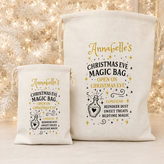 Two cream canvas drawstring bags with gold and black printed holiday text and illustration, shown on a light surface with blurred warm white Christmas lights in the background.