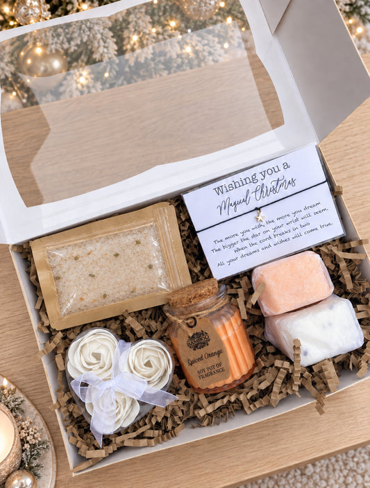An open Secret Santa gift box filled with spa and self-care items on a wooden surface, including a candle jar, two shower steamers, a packet of bath salts, rolled bath soap, and a handwritten gift message card.