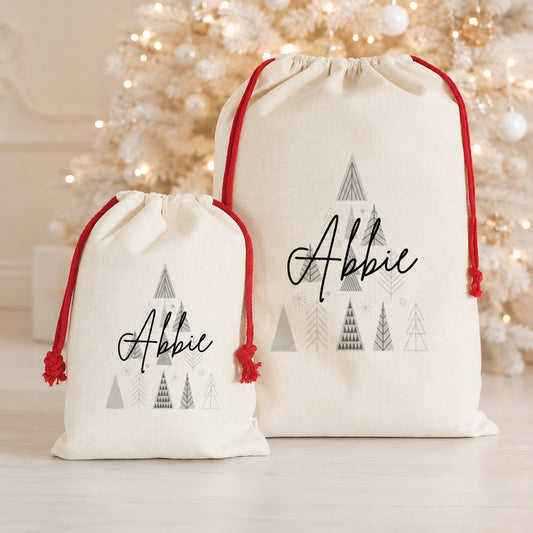 Two personalized cream canvas Santa bags with red drawstrings and a gray Christmas tree design, displayed in front of a softly blurred festive Christmas tree.