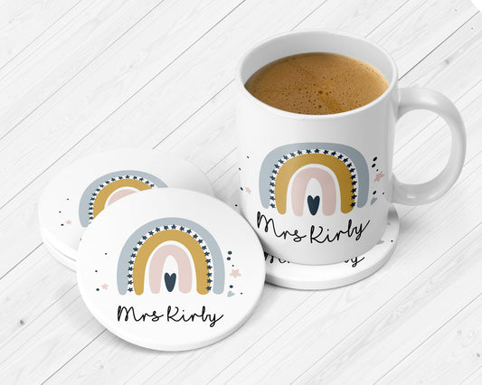 White ceramic mug and coasters set personalised with the name “Mrs Kirby.” Features a whimsical rainbow design in muted pastel tones of blue, gold, and blush pink, with surrounding star and dot accents.